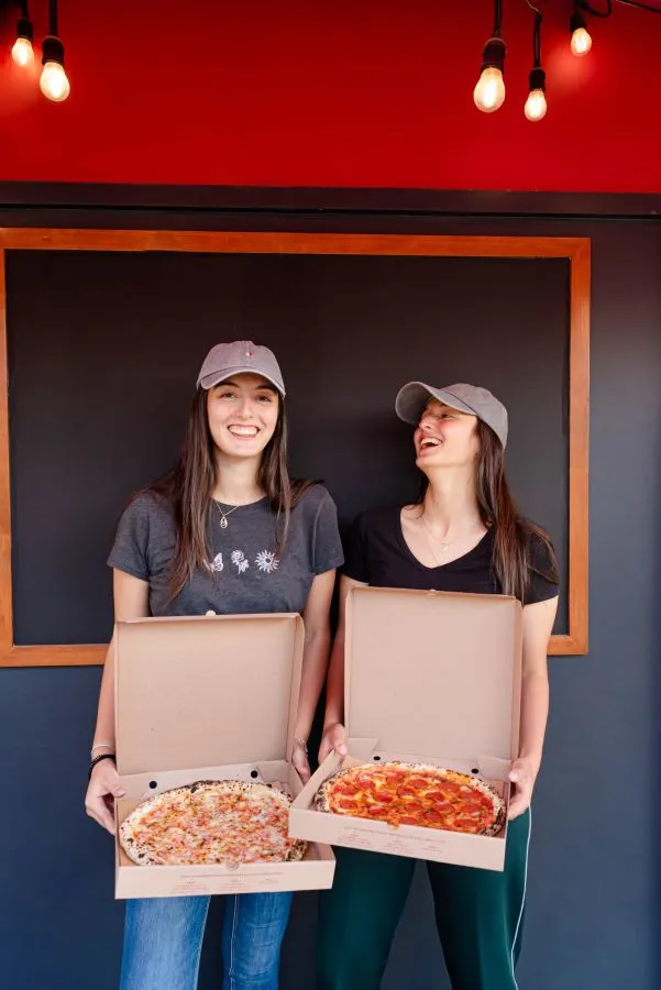 Dos chicas sosteniendo dos cajas de pizzas.